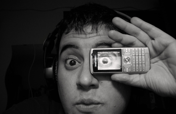A person wearing headphones and holding a small, old mobile phone in front of their left eye showing a photograph of their left eye, while looking surprised at something