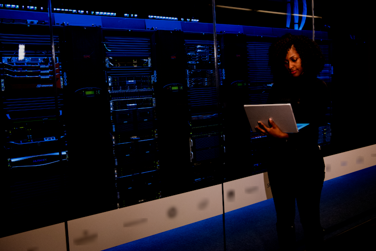 A Black woman with an open laptop, walking past a row of server racks