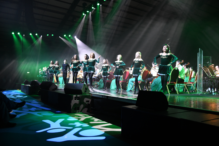 A line of Irish dancers on a stage, all dressed in green, dancing in front of a band
