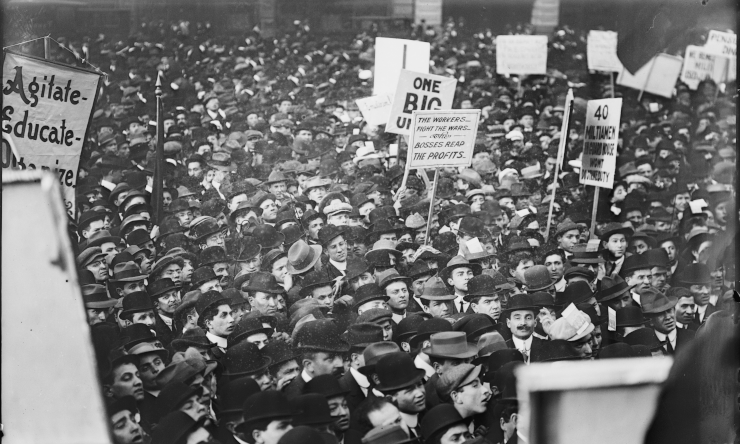 A large audience, some members holding signs saying such slogans as "Agitate, Educate, Organize," "One Big Union," "The Workers Fight the Wars While Bosses Reap the Profits," and so forth