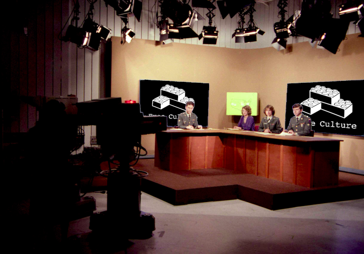 A news set with the logo representing Free Culture, featuring a sketch of three definitely generic modular brick toys stacked as a corner, showing on screens behind the anchors