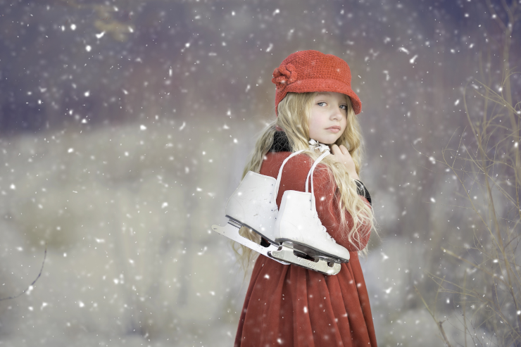 A girl in a red cloak and hat carrying white ice skates over her shoulder in a snowy outdoor area
